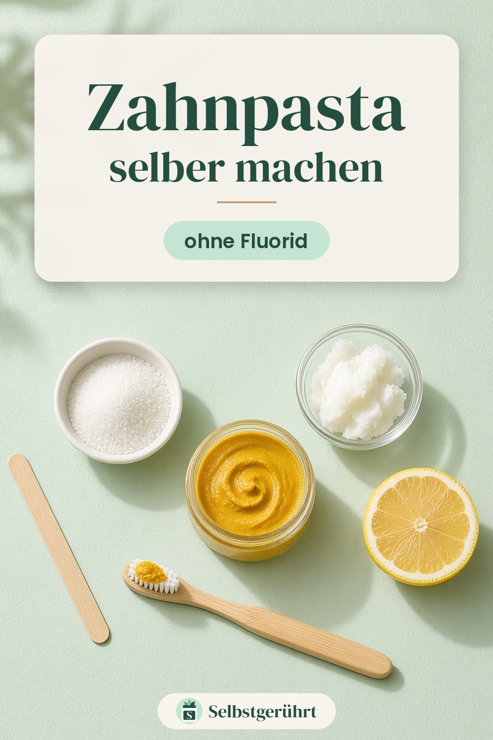 Flatlay auf mintgrünem Untergrund: offener Glastiegel mit gelblicher Zahnpasta, daneben eine Bambus-Zahnbürste mit einem Klecks Paste, ein flacher Holzspatel, ein Schälchen Xylit, ein Schälchen Kokosöl und eine halbe Zitrone. Oben steht „Zahnpasta selber machen – ohne Fluorid“.